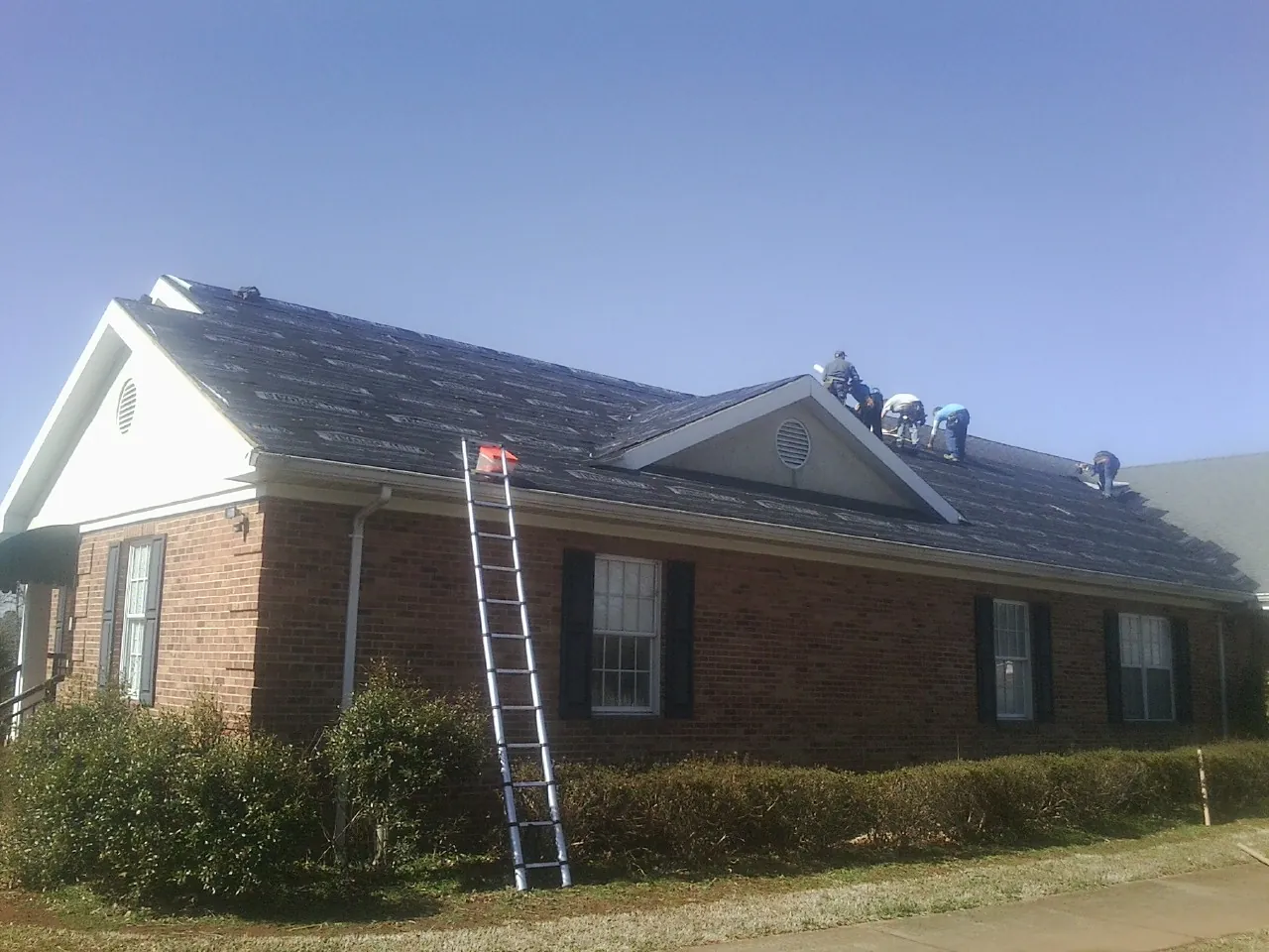 Crew tearing shingles off a brick ranch roof with a ladder set — Emergency Roof Repair work by our crew in Clinton